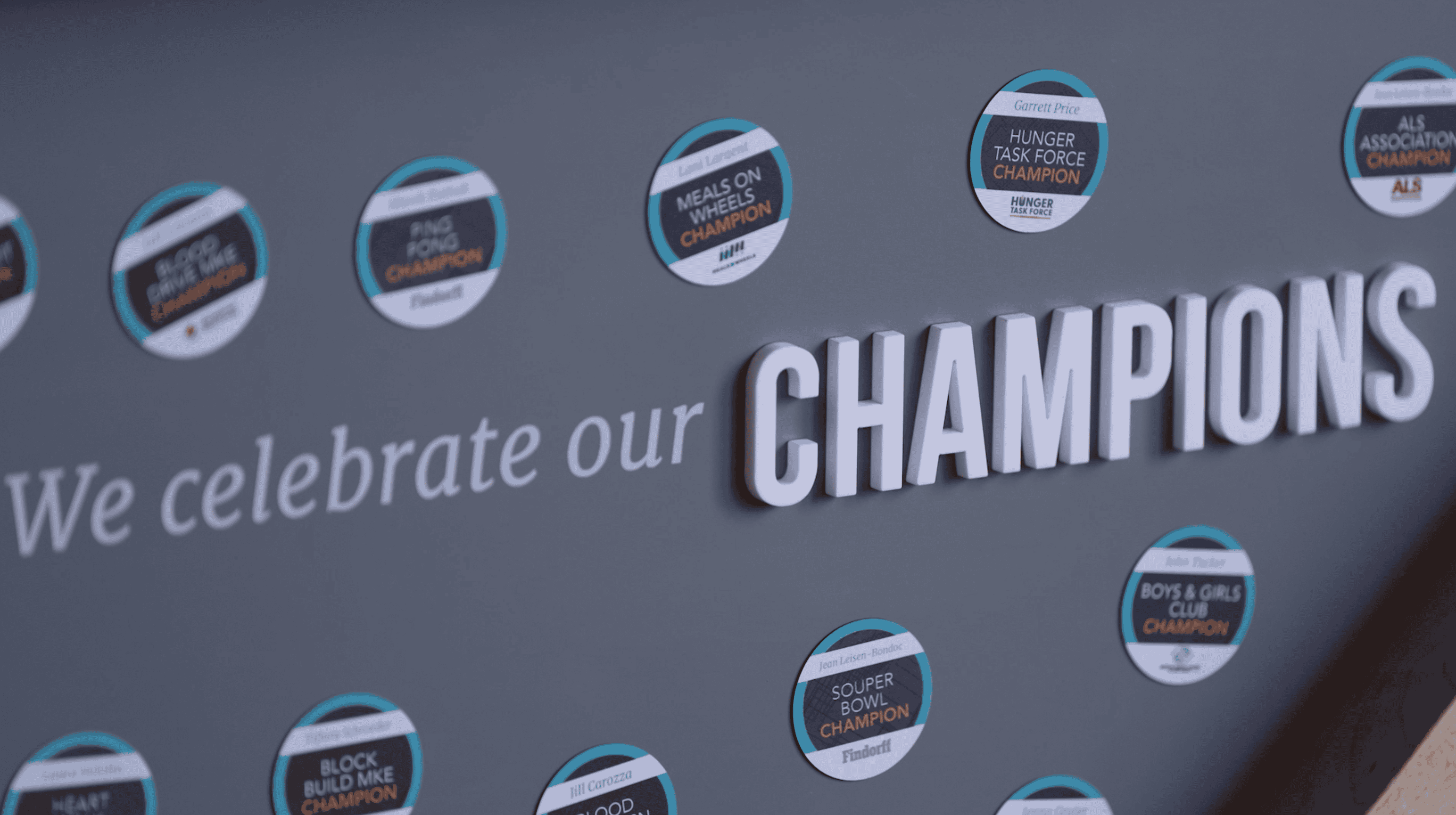 A display wall with the text "We celebrate our CHAMPIONS" in bold white letters on a gray background. Surrounding the text are circular badges, each representing different champion titles, such as "Boys & Girls Club Champion" and "Kids Building WI Champion."